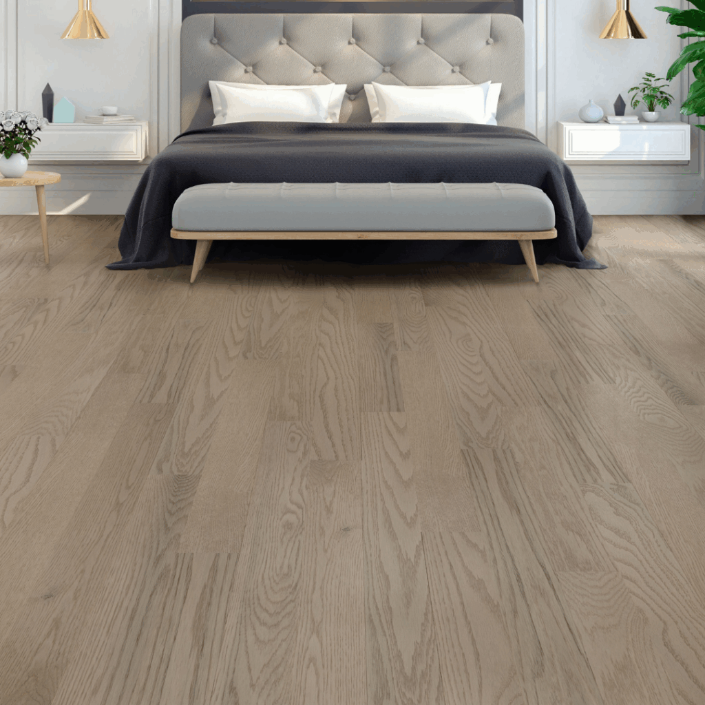 Modern bedroom with a large bed, upholstered headboard, gray blanket, white bench, nightstands, and light wood flooring. Plants and wall lamps add decorative accents.