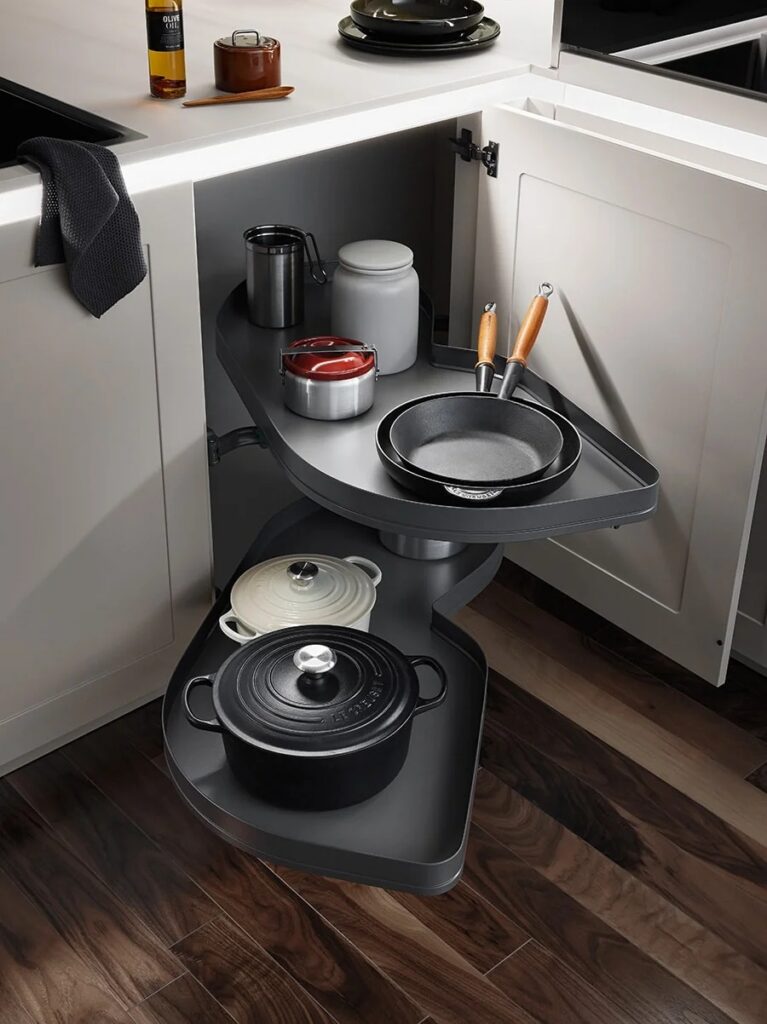 A corner kitchen cabinet with two pull-out shelves holding cookware, including cast iron pots, pans, and utensils, on a wood floor.