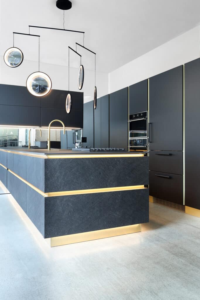 Modern kitchen with dark matte cabinets, built-in appliances, a large island with gold accents, and round pendant lights hanging above.