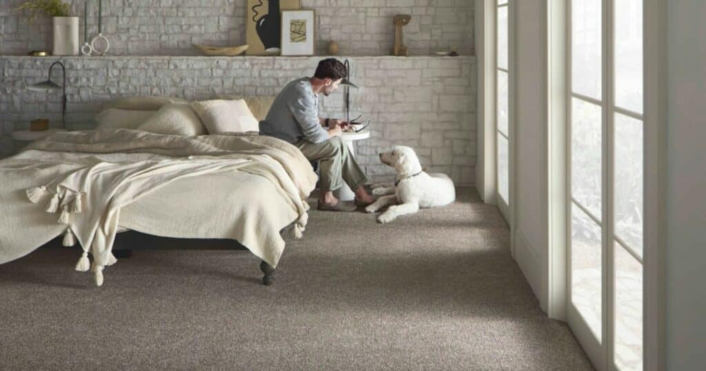A man is sitting on a bed in a bedroom, surrounded by the latest carpet innovations that promote health and hygiene.